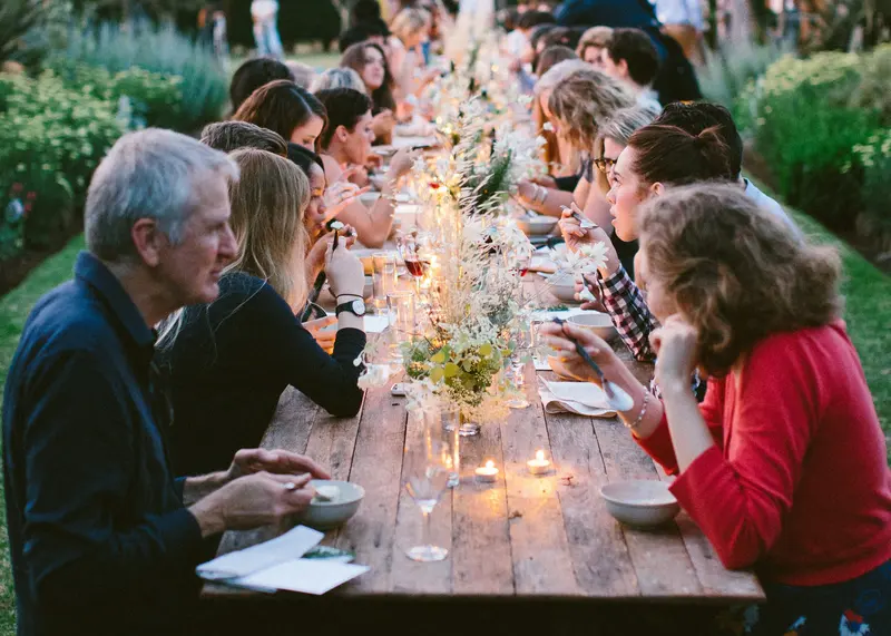 Invitados conversan en una mesa larga al aire libre, entre plantas y con velas encendidas